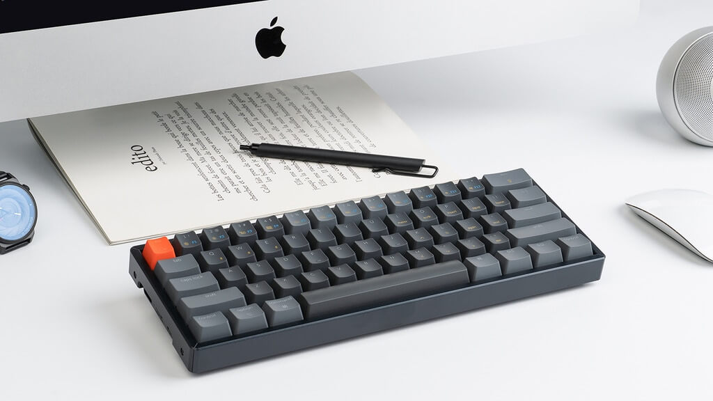 A black and grey keyboard, resting on a table beneath a Mac monitor.